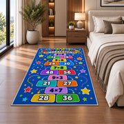 Multiplication Hopscotch Rug shown beside a bed with neutral bedding